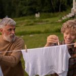 Elderly man and teenage boy joyfully hanging laundry in a sunny garden setting.